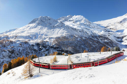 A red train is passing the train tracks with tight 180° curve at high Alp Grum. The Piz Palu peak is at the background.